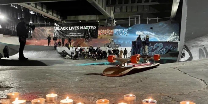 Crowd standing in the bowl of the burnside skate park listening to a speaker. Candles and a skateboard shown at the front of the photo