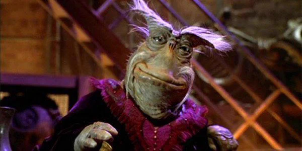 Dominar Rygel XVI from Farscape. He is a small alien creature with wrinkly green skin and a long face. His features resemble a fish or toad and he has protruding eyebrows with grey hair. He wears ornate clothing and is smiling at the camera.