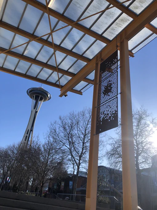 A rectangular overhead shelter supported by pillars. Between them are three rusted metal panels punched out with Penrose tiling of small diamonds. The sky is blue and the Space Needle stands tall in the background.