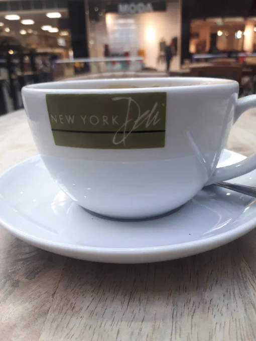 Photo of a white cup and saucer on a stripped wooden table. At the top of the cup is a khaki rectangle with the words New York Deli in white text, underlined in black. In the background is a blurred image of a clothes shop called MODA.