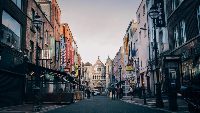 Anne Street leads down to St Anne’s Church, a large and intricate tan building. On either side of the street are numerous shops including restaurants, cafes, and a barber shop. Scattered people are walking along the street. Above the buildings, the sky is a light grey. 