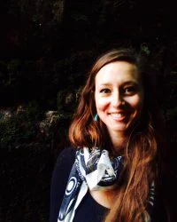 A portrait of a person standing outside against a dark background. She's wearing a black shirt and a patterned scarf, and she's smiling at the camera.