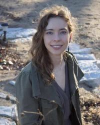 A portrait of a person standing outside. She is wearing a green jacket and brown shirt, and she's smiling at the camera.