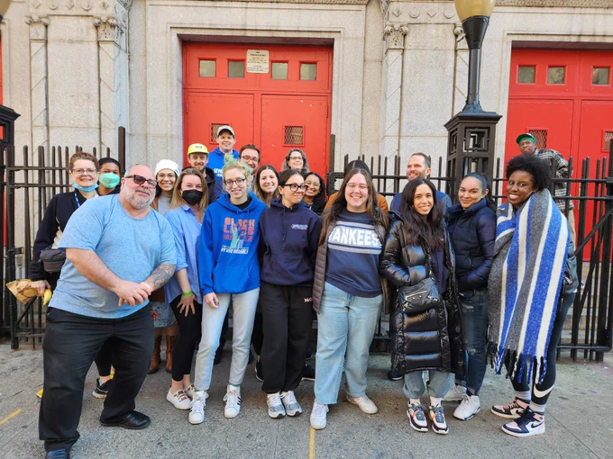A group of teachers gathers outside the red doors of a high school, smiling for the camera.