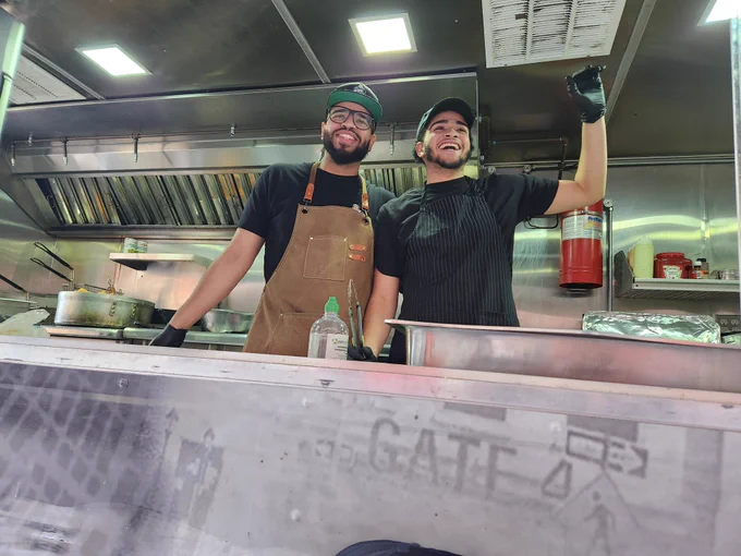 Two men wearing baseball caps and aprons smile and wave from the inside of a food truck.