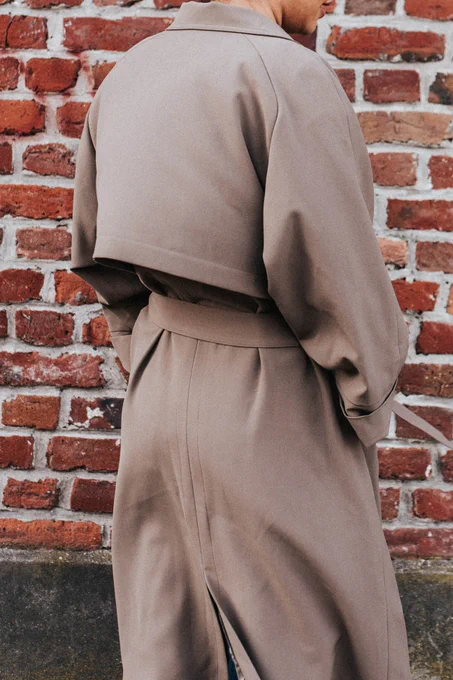 La mannequin est de dos face à un mur de briques rouges. Elle porte le trench couleur tabac.

Trench double boutonnage, poches à large passepoil. Manches longueur 7/8ème avec grand revers. Col droit. Poche intérieure passepoilée. Bas-volets au dos et devant. Entièrement doublé. Fente milieu dos. Ceinture à nouer qui se glisse dans les passants côtés. 