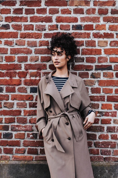 La mannequin est appuyée contre un mur de briques rouges. Elle porte le trench couleur tabac avec en dessous un body hugo rayé façon marinière.

Trench double boutonnage, poches à large passepoil. Manches longueur 7/8ème avec grand revers. Col droit. Poche intérieure passepoilée. Bas-volets au dos et devant. Entièrement doublé. Fente milieu dos. Ceinture à nouer qui se glisse dans les passants côtés. 