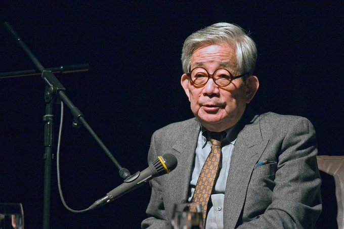 Kenzaburō Ōe speaking at an event. He is wearing a grey suit and bronze tie. He has round glasses.