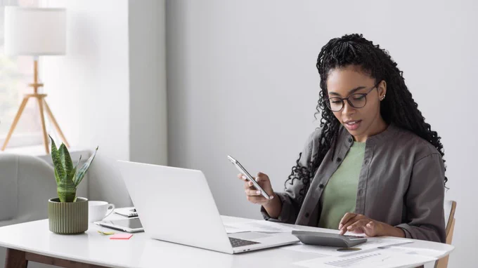 A woman sits at her desk in front of an open laptop holding her phone.
