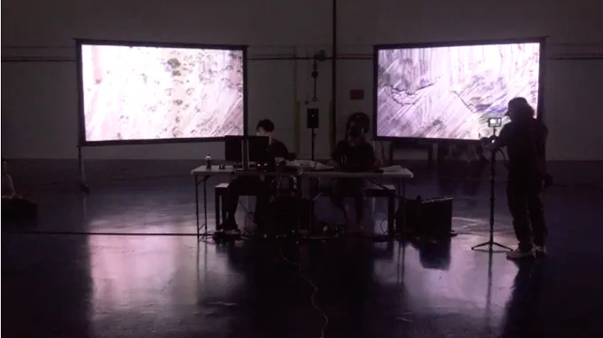 a screenshot of the performance with 2 large panels behind 2 performers and a camera person on the right just in front of the right panel