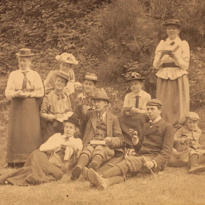 A group of students are outdoors writing in their notebooks. The Woodwardian Professor is pointing at something. The group are all wearing hats. Some are holding geological hammers. 