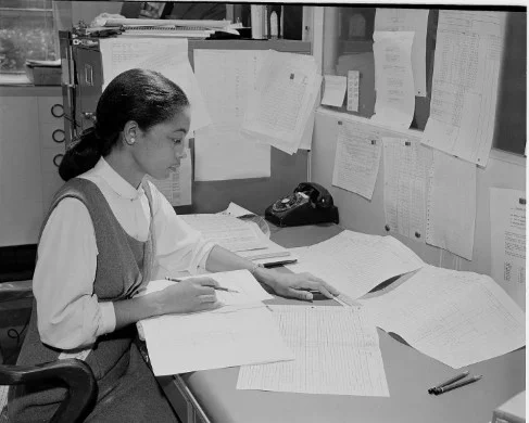 Black and white image of Inez Hazel working at a desk and review various pages of paper.