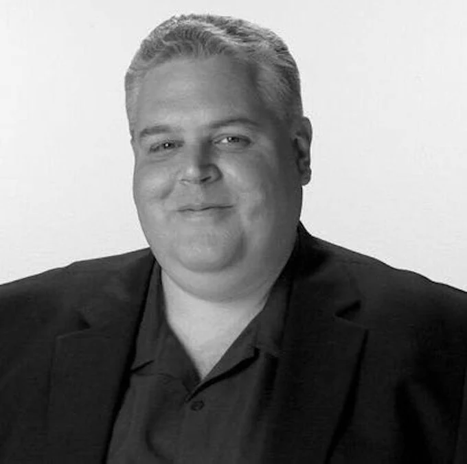 A black and white headshot of me, a devastatingly handsome white dude with short white and gray hair, smiling and wearing a suit jacket with a collared shirt and no tie.