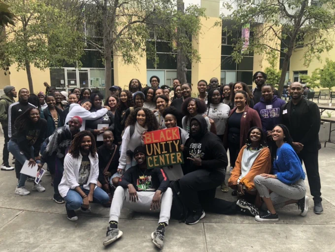 About 40 students pose for a group photo outside of the Student Services Buildinig with one person holding a sign with the text Black Unity Center
