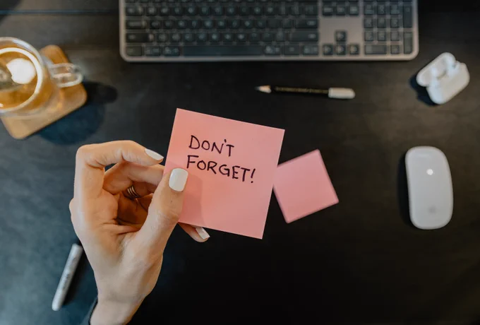 A light-skinned hands with painted white fingernails holds a sticky note with the words: "Don't forget!" written on it.