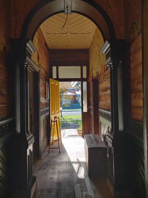 Looking up the unrepaired hallway towards the open front door, with morning sun light coming and highlighting the yellow painted door. Some bits and pieces of renovation stuff are cluttering up the hall. 