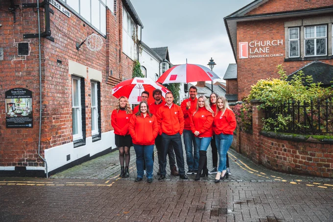 The Red Box Web Design Ltd Team wearing Red coloured sweatshirts with umbrellas in Church Lane in Stafford, outside the Soup Kitchen.