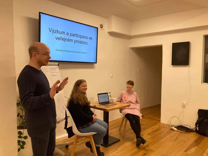 Dominika Potužáková (vpravo) a Tereza Fasurová (uprostřed) sedí před prezentací na velké televizi. Martin Kopta (stojí vlevo) dlaněmi otočenými k publiku vysílá pozitivní energii.