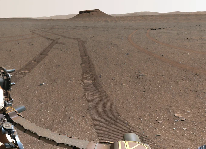Panoramic photo from NASA’s Mars Perseverance Rover shows wheel tracks across a vast, flat, desert landscape. Along the tracks, which recede into the distance in a zig-zag pattern, are 10 small sample tubes placed on the surface by the rover.  Part of the rover’s robotic arm is visible at lower left. The horizon is dominated by a large mesa-like feature. Older wheel tracks, also created by Perseverance, can be seen on the right, appearing somewhat fainter than the fresh tracks on the left.