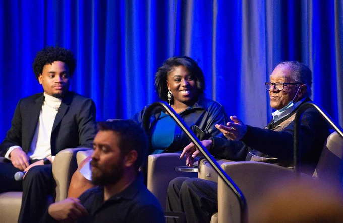 Two CSU students and Bobby Seale sitting on chairs on stage. 