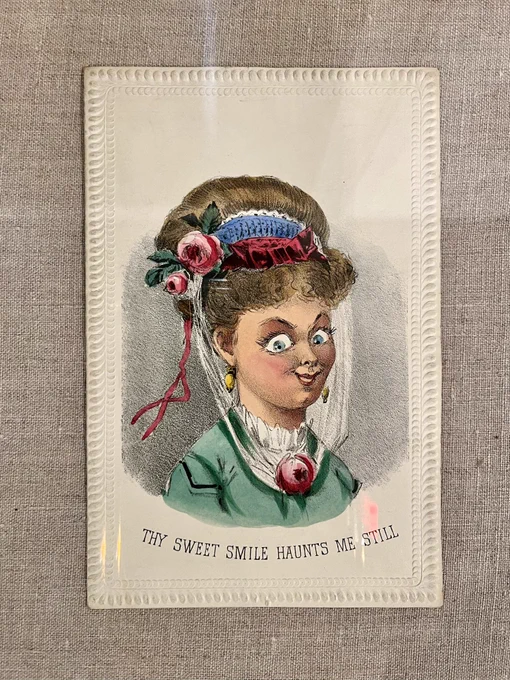 Valentine card featuring an illustrated portrait of an elaborately dressed woman with creepy eyes and arched eyebrows who is smiling at the viewer. Caption below reads, “Thy sweet smile haunts me still”.