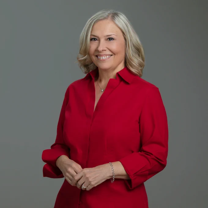 Swing Time! host Jan Eberle poses for a portraiture, medium length blonde hair, with bright smile in a red dress. She wears a diamond  solitaire necklace, a bracelet on her left wrist, and a diamond ring is seen on her third finger, left hand. 