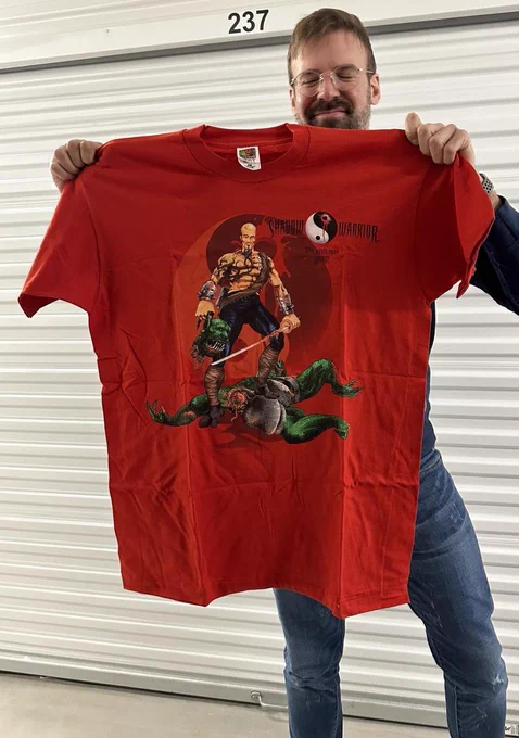 Apogee's Executive Producer, Jeron Moore, holding a vintage blood-red t-shirt featuring SHADOW WARRIOR branding and Lo-Wang, decapitating his enemy.