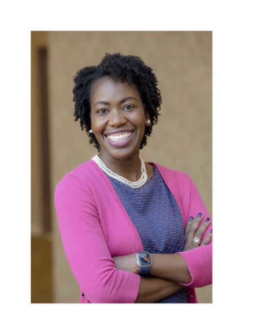 Black woman wearing a pink sweater, a blue shirt with pink dots, and pearl earrings and necklace.