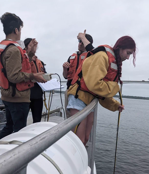 A research student on a boat deck lowers a water monitoring instrument into the water. Three other people are with her on the boat's deck.