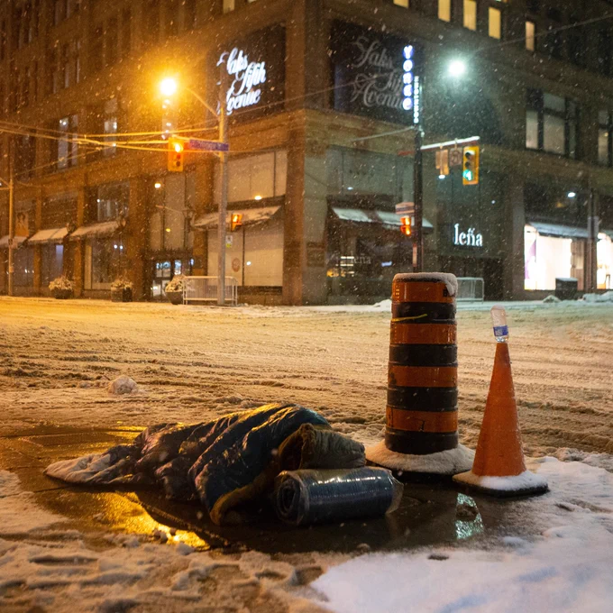 A photo, taken by Chris Young, of a man sleeping rough outdoors on a snowy ground in front of a Saks Fifth Avenue. 