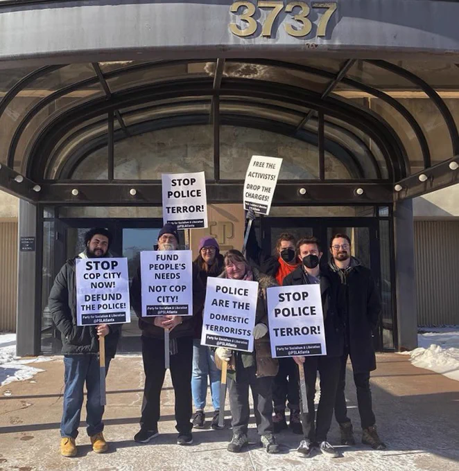 Group of community members holding signs and standing in front of an office building that houses Cushman & Wakefield. Signs read “Stop cop city now! Defund the police” “Fund people’s needs not cop city!” “police are domestic terrorists” “Stop police terror!” “Free the activists! Drop the charges!