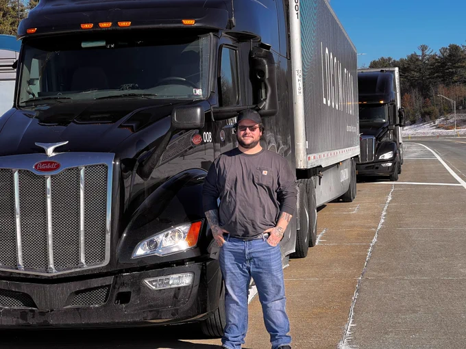 Zac Gabert, a Locomation Autonomous Truck Operator, stands in front of a black two-truck commercial motor vehicle convoy with Locomation branding. 