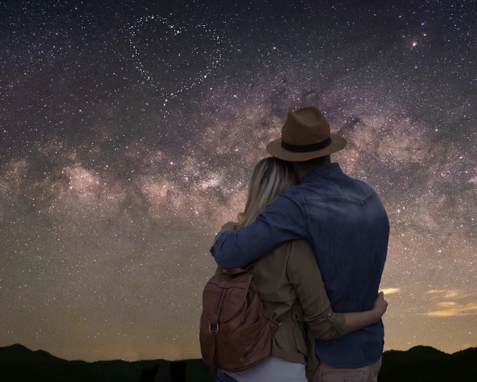A couple embraces underneath a starlit night sky. Above the Milky Way, there is an asterism in the shape of a heart.