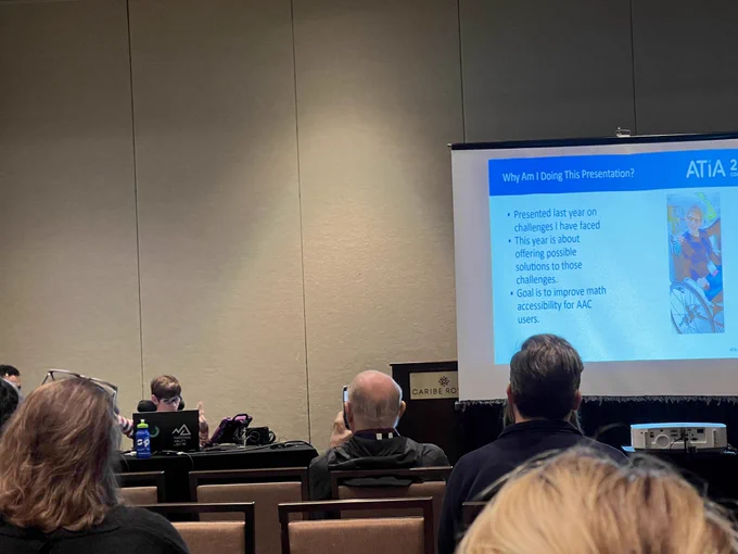 Photo of presenter LJ - a teenager with brown heart and eye glasses sitting by his computer and AAC device presenting. Some audience members are watching/listening to the presentation. The screen projection of the slide show is located to the front left of the photo.