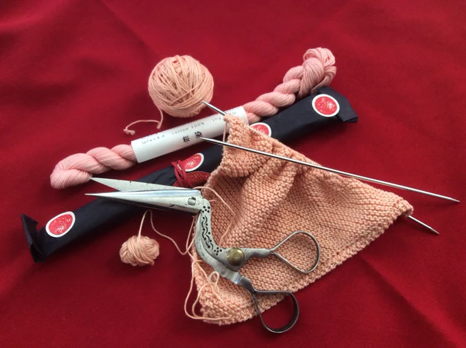 A ball of pale pink yarn, a twisted hank of pink yarn, a black tissue wrapped pack which contains pink yarn, and some knitting - a triangle - on dpns which is in pink yarn. There’s also a pair of slightly crudely made stork scissors. Red background. Which is a napkin, I think a friend nicked this from somewhere.