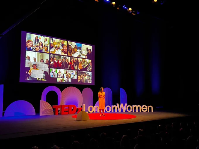 Anneka on stage on a big red circular carpet, wearing a yellow dress. Lots of images of women on screen behind her.