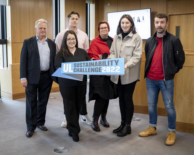 A photograph of a group of six people taken in a room with wood paneled walls and grey carpet. They are all smiling for the camera and holding a blue sign which says "UL Sustainability Challenge 2022"