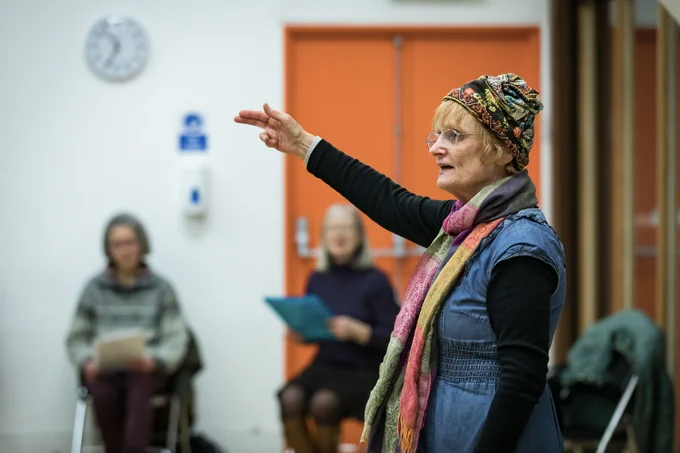 A person wearing a denim dress and colourful patterned scarf and hat stands in front of a group of seated adults. The person standing has one arm raised and is pointing forwards. darts musician Janet Wood leads a Breathe & Connect session for adults with Long Covid at The Point in Doncaster.