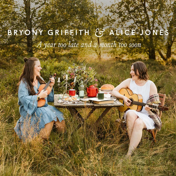 Album cover for 'A Year Too Late and a Month Too Soon' by Bryony Griffith and Alice Jones. There is a photo of Bryony and Alice sat at a table outside, playing fiddle and guitar and laughing and joking. The table is covered with flowers, candles, a teapot, a book and other bits and pieces. There are trees in the background behind them.