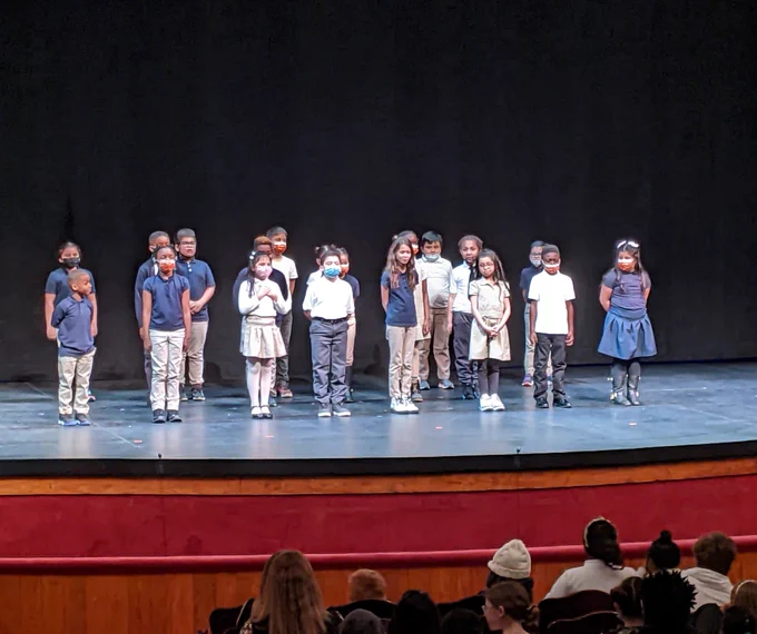 A class of upper elementary school students wearing navy blue and beige uniforms stand onstage at Ford's Theatre while delivering a speech during the 2023 Lincoln Oratory Festival.