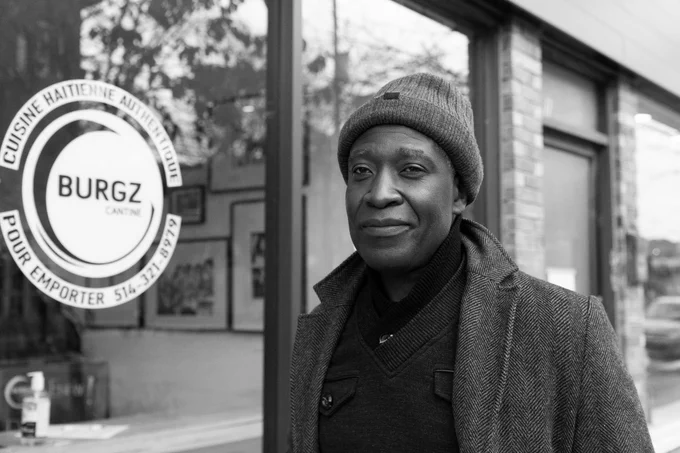 A man in a peacoat and toque stands outside a shop window that says, 'Burgz'. He is slightly smiling. Photo by Stacy Lee.