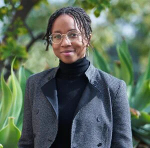 Black woman in glasses wearing a dark gray jacket over a black turtleneck, with green leaves in the background.