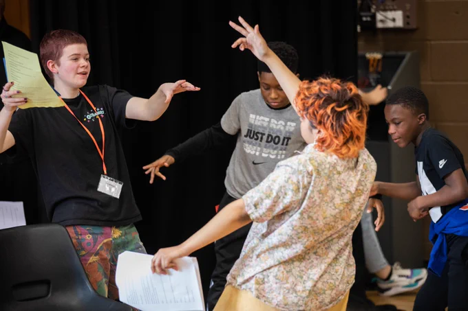 Four young people. The two in the foreground face each and hold sheets of paper with one hand whilst they mimic an explosion with the other. The other two in the background look down. Their poses are dynamic.