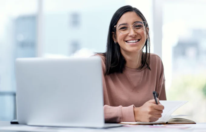 A woman with glasses smiling at the camera sits at a laptop computer while holding a pen and writing in a notebook.