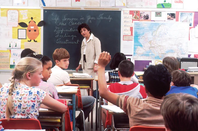 The photo shows a classroom with pupils, a teacher is standing at the blackboard talking to the class.