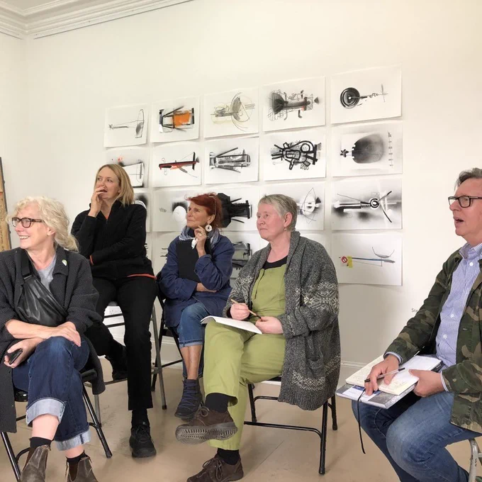 Photograph supplied by Off The Rails Arthouse. Pictured 5 people sitting on chairs in an art studio, with a series of mostly monochromatic artworks on the wall behind them. Some of the people have sketchbooks and pencils and all are involved in a group discussion or talk.