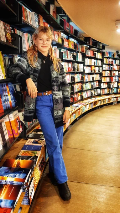 Romy, wearing a gray cardigan, a black shirt, and blue pants with a brown belt, casually leaning against our Amsterdam store's Fiction section on the second floor.