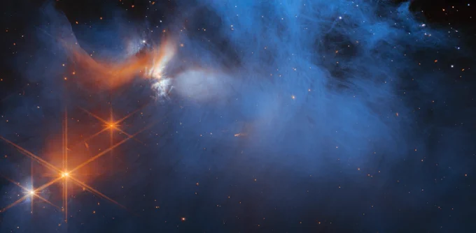 A JWST image of the central region of the Chamaeleon I dark molecular cloud, which resides 630 light-years away. This image primarily shows blue smoky wisps on a dark background. The left top side additionally features orange and white wisps. Just below them are four bright points of light. Three are orange and one is a mix of white and orange. Each of these points have JWST’s signature 8-point diffraction spikes emanating around them in long, thick orange lines, so that they look like huge snowflakes. Scattered throughout the image are distant stars or galaxies in shades of red, orange and blue, seen as tiny blobs.
