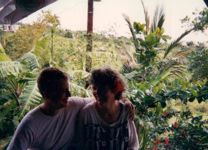 Leslie Feinberg & Minnie Bruce Pratt in Vieques, January 1993, in t-shirts, Leslie's arm around MB shoulder, MB with red hibiscus behind one ear, wild green vegetation behind.
