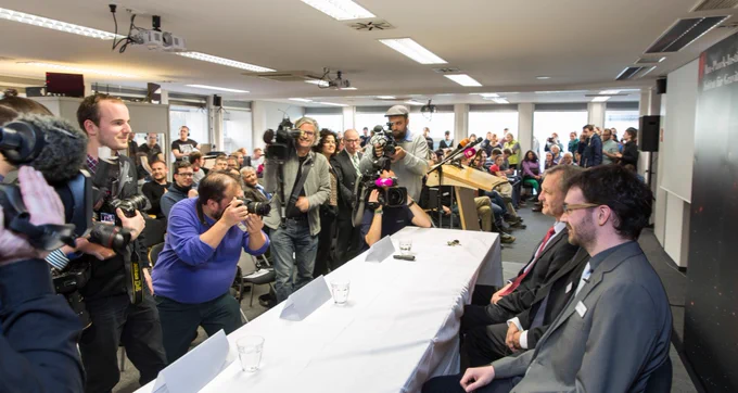 In a large room, on the left reporters and camera persons film two persons in suits on the left. In the background a large number of people and a lectern.